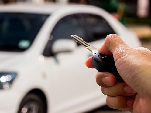 a imagem ilustra uma mão segurando a chave de um carro e ao fundo um carro branco